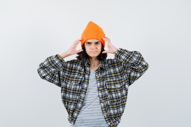 Young woman holding hand near head in orange hat checkered shirt looking displeased