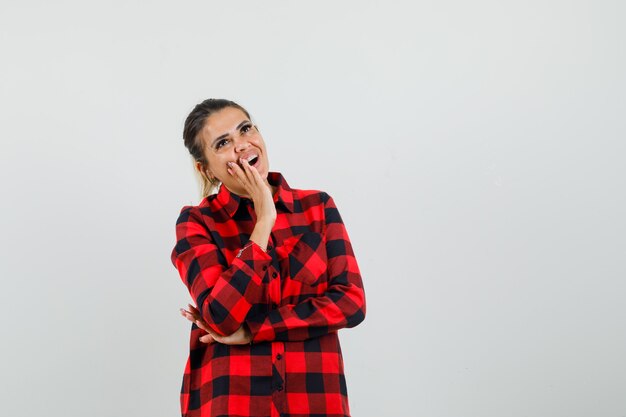 Young woman holding hand near mouth in checked shirt and looking happy. front view.