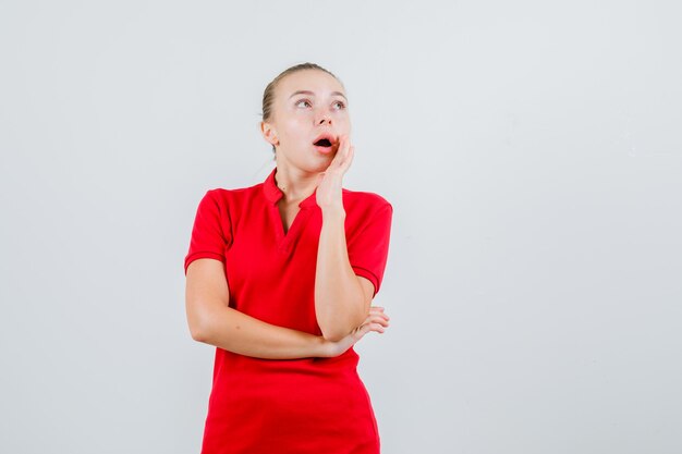 Young woman holding hand near mouth in red t-shirt and looking amazed