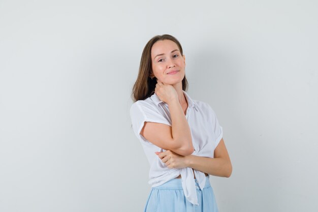 Young woman holding hand on neck and smiling in white blouse and light blue skirt and looking charming