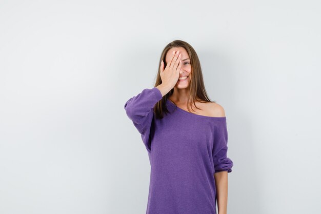 Young woman holding hand on one eye in violet shirt and looking happy. front view.