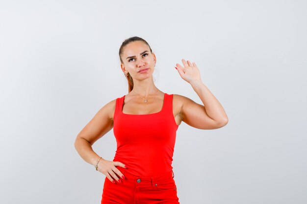 Young woman holding hand on waist, showing stop gesture in red tank top, pants and looking confident , front view.