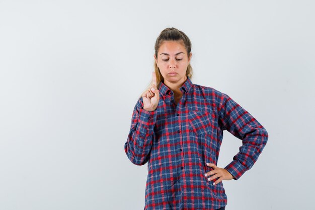 Young woman holding hand on waist while raising index finger in eureka gesture in checked shirt and looking serious
