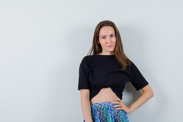 Young woman holding hand on waist while smiling in black t-shirt and blue skirt and looking confident