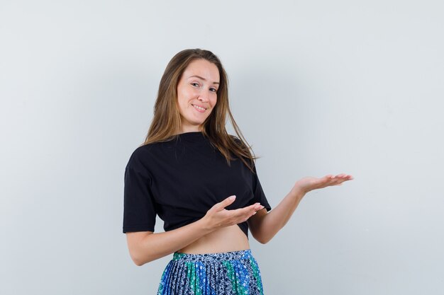 Young woman holding hands as presenting something in black t-shirt and blue skirt and looking happy