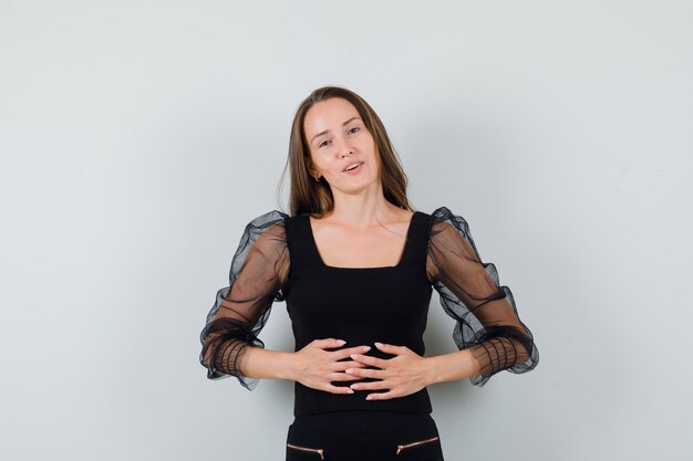 Young woman holding hands on belly in black blouse and black pants and looking optimistic 