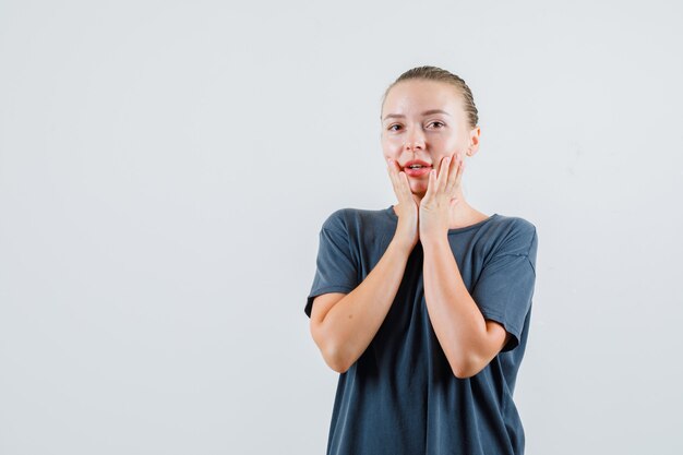 Young woman holding hands on cheeks in gray t-shirt and looking pretty