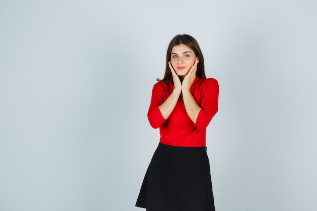 Young woman holding hands on cheeks in red blouse, black skirt and looking sanguine