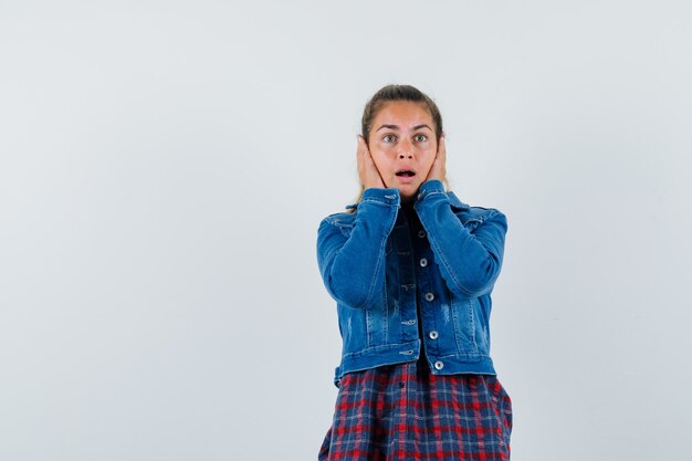 Young woman holding hands on cheeks in shirt, jacket and looking anxious , front view.