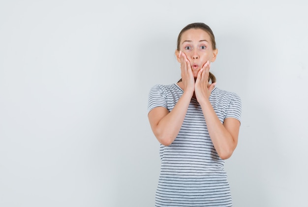 Young woman holding hands on cheeks in t-shirt and looking surprised , front view.