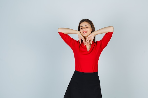 Young woman holding hands under chin, keeping eyes closed in red blouse