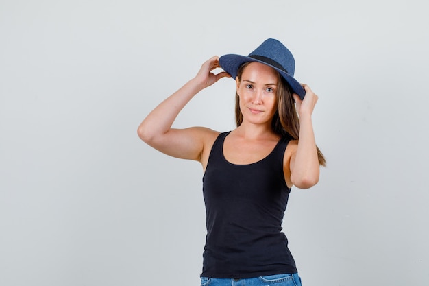 Young woman holding hands on hat and smiling in singlet, shorts front view.