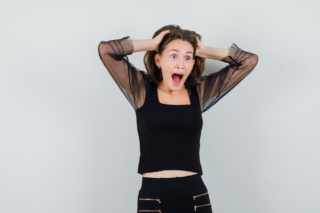 Free photo young woman holding hands on head in black blouse and black pants and looking surprised