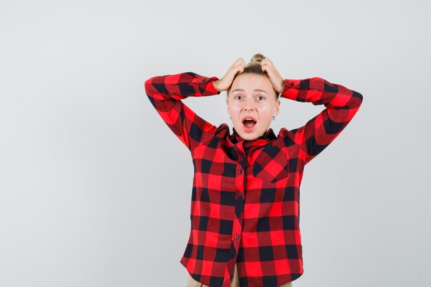 Young woman holding hands on head in checked shirt, pants and looking forgetful