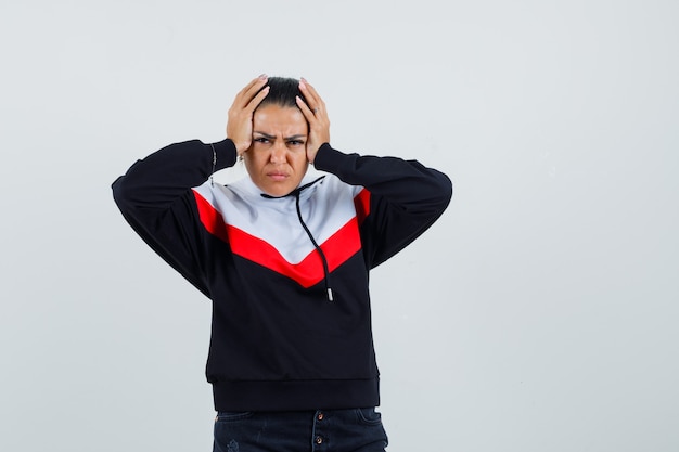 Giovane donna che tiene le mani sulla testa, avendo mal di testa in maglione e jeans neri e guardando infastidito. vista frontale.