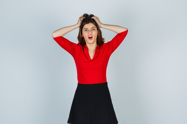 Young woman holding hands on head in red blouse, black skirt and looking annoyed
