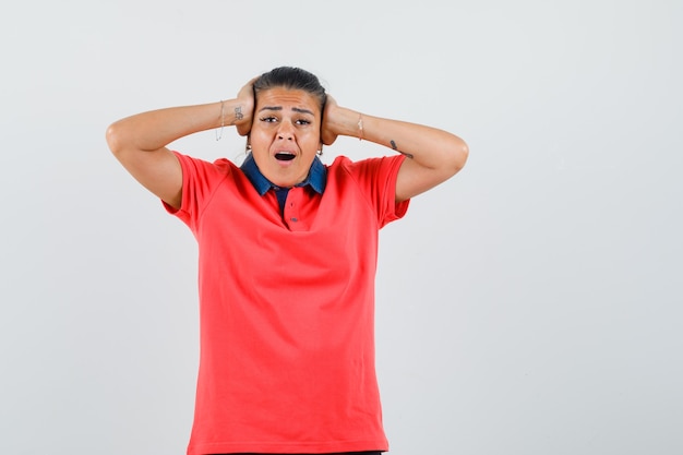 Young woman holding hands on head, screaming in red t-shirt and looking annoyed. front view.