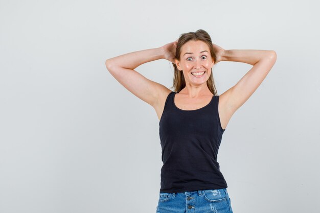 Young woman holding hands behind head in singlet, shorts and looking funny