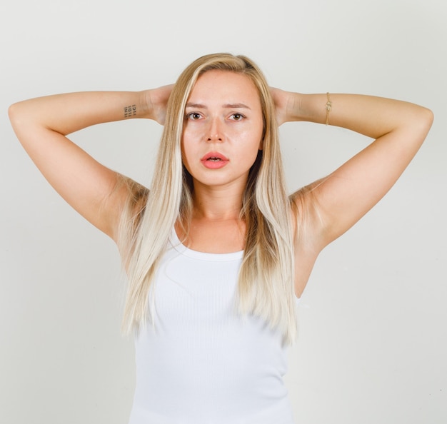 Free photo young woman holding hands behind head in white singlet and looking pensive.