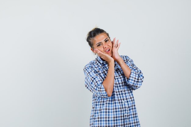 Young woman holding hands on her cheeks in checkered shirt and looking uncomfortable.