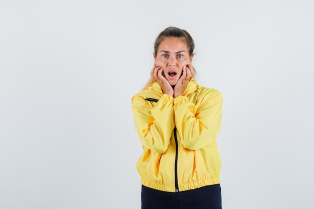 Young woman holding hands on her dropped jaw in yellow raincoat and looking nervous