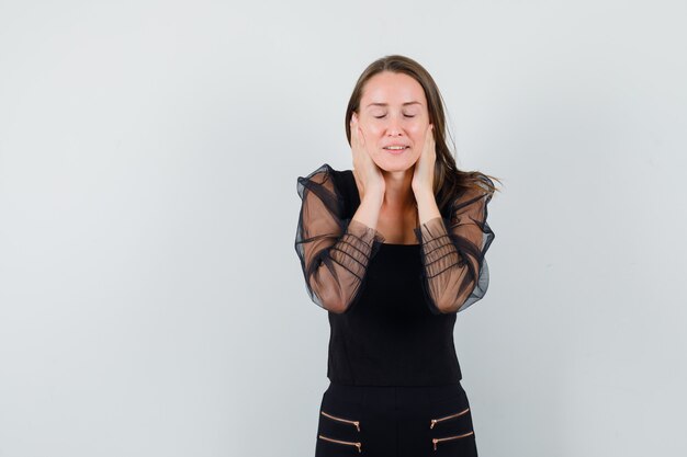 Young woman holding hands on her ears while closing eyes in black blouse and looking calm. front view. space for text