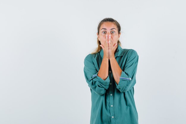 Young woman holding hands on her mouth while opening eyes widely in blue shirt and looking terrified