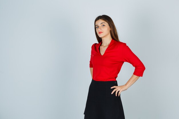Молодая женщина, держащая руки на бедрах в красной блузке, черной юбке и уверенная в себе