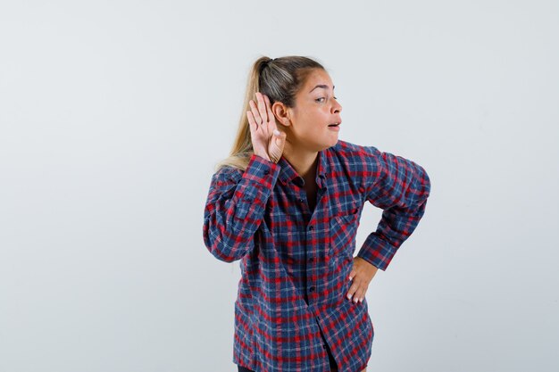 Giovane donna che tiene le mani vicino all'orecchio per sentire qualcosa in camicia controllata e guardando concentrato