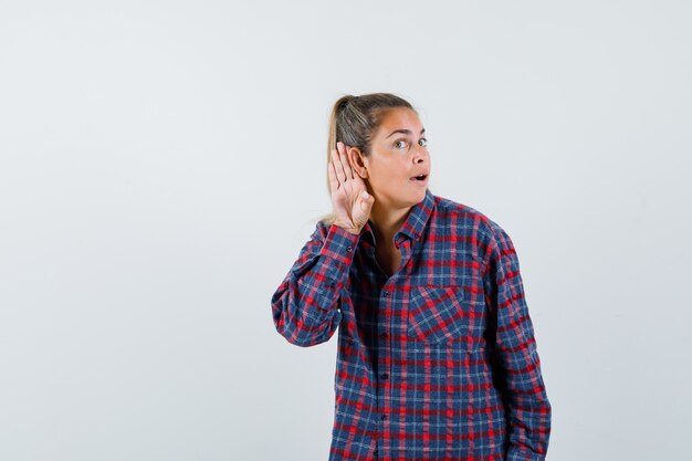 Young woman holding hands near ear to hear something in checked shirt and looking happy