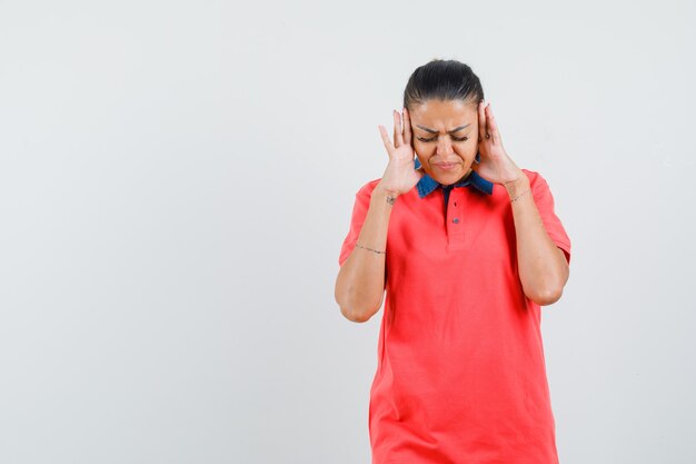 Young woman holding hands near head, having headache in red t-shirt and looking exhausted. front view.