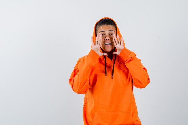 Young woman holding hands near mouth as calling someone in orange hoodie and looking focused 