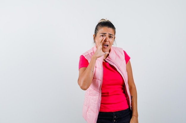 Young woman holding hands near mouth as calling someone in pink t-shirt and jacket and looking focused 