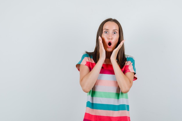 Young woman holding hands near mouth in t-shirt and looking shocked , front view.