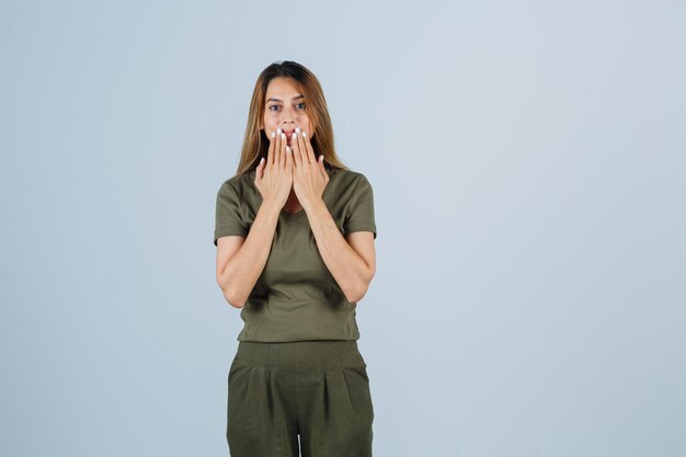 Young woman holding hands near mouth in t-shirt, pants and looking surprised. front view.