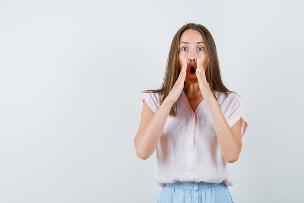 Young woman holding hands near mouth in t-shirt, skirt and looking scared. front view.