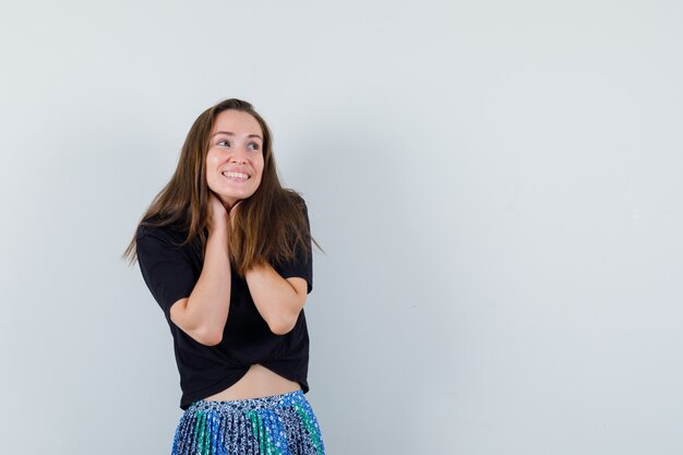 Young woman holding hands on neck and looking away in black t-shirt and blue skirt and looking happy