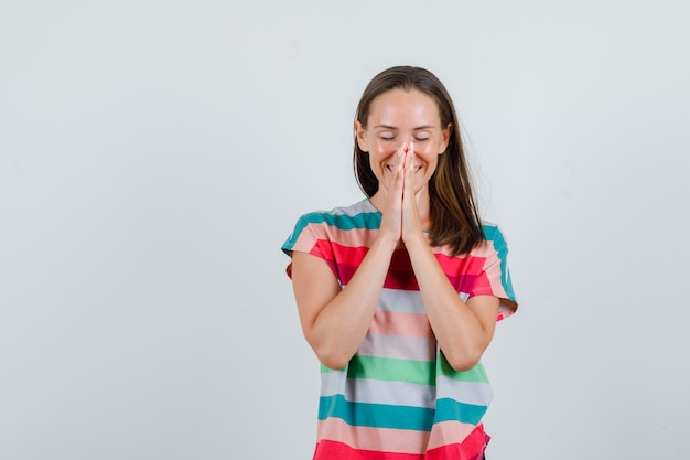 Young woman holding hands in praying gesture in t-shirt and looking grateful , front view.