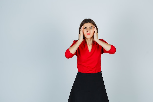 Young woman holding hands on temples in red blouse, black skirt and looking worried