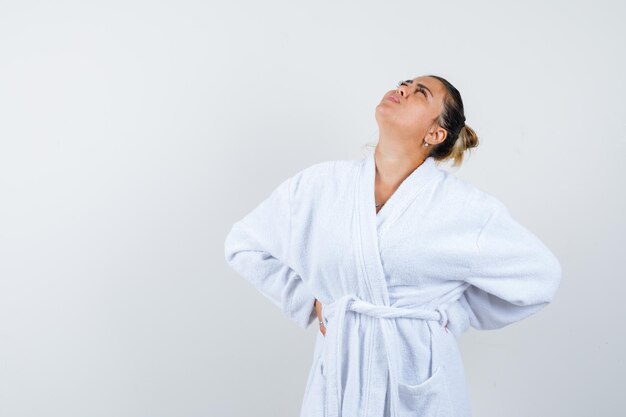 Young woman holding hands behind waist, looking above in white bathrobe and looking pretty