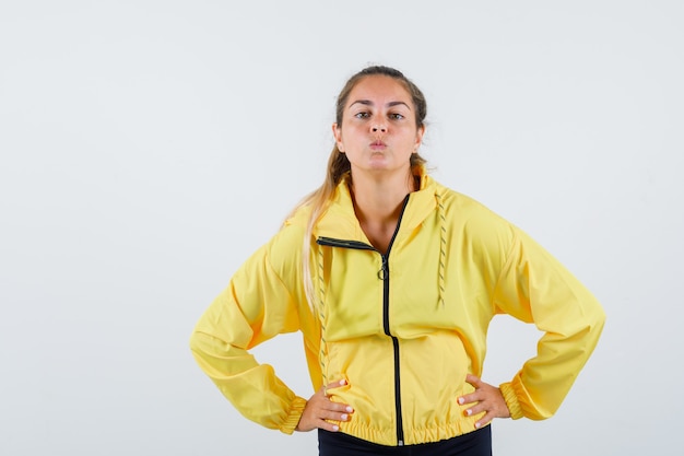 Young woman holding hands on waist while pouting lips in yellow raincoat front view.