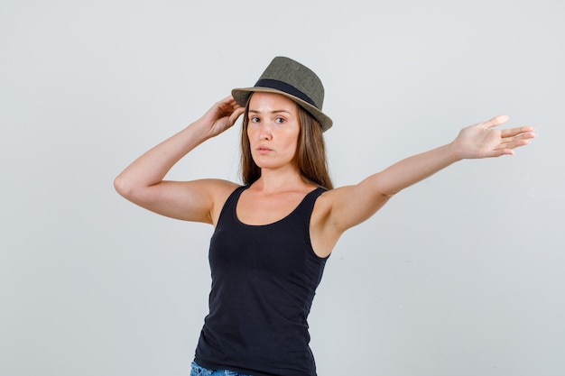 Young woman holding hat while stretching arm in singlet, shorts front view.