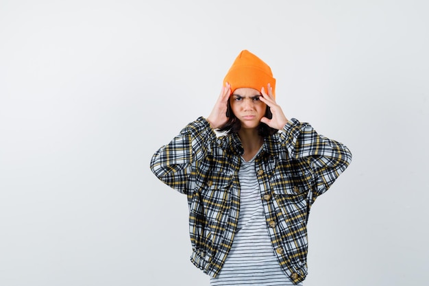 Young woman holding head with fingers in orange hat checkered shirt looking painful