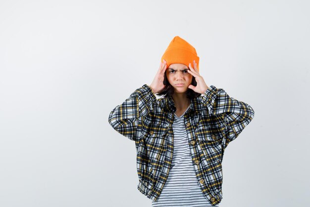 痛みを伴うオレンジ色の帽子の市松模様のシャツで指で頭を保持している若い女性