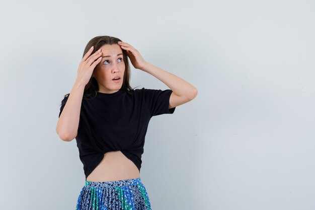 Young woman holding head with hands, looking away in black t-shirt and blue skirt and looking serious