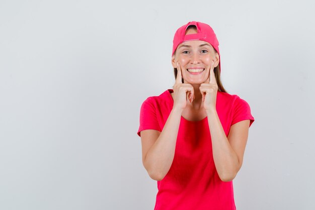 Young woman holding index fingers on cheeks in pink t-shirt, cap and looking glad. front view.