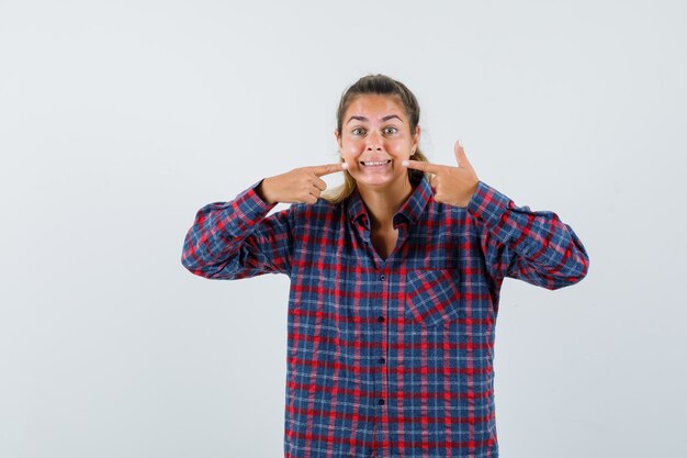 Young woman holding index fingers near mouth, forcing a smile in checked shirt and looking happy