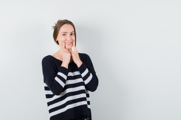 Young woman holding index fingers near mouth and smiling in striped knitwear and black pants and looking happy