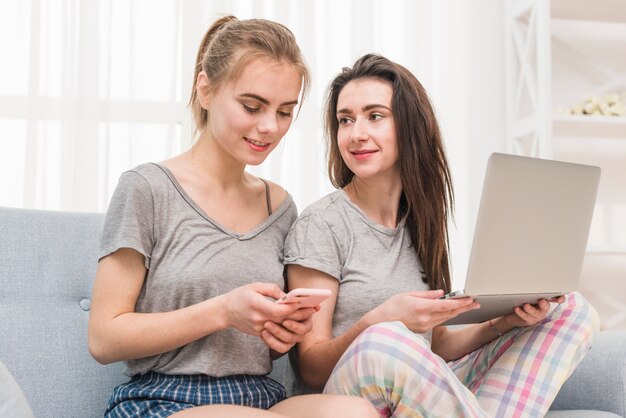 Young woman holding laptop in hand looking at her girlfriend using mobile phone at home