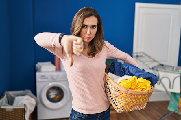 Young woman holding laundry basket looking unhappy and angry showing rejection and negative with thumbs down gesture. bad expression.